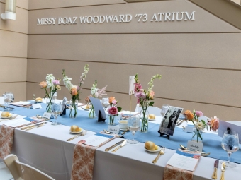 A long dining table set with white linens, blue runners, floral arrangements, and menus sits beneath a wall inscribed with "MISSY BOAZ WOODWARD '73 ATRIUM".