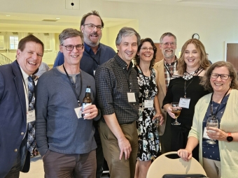 Seven smiling alumni wearing Davidson College name tags pose together at an indoor reception while holding drinks.