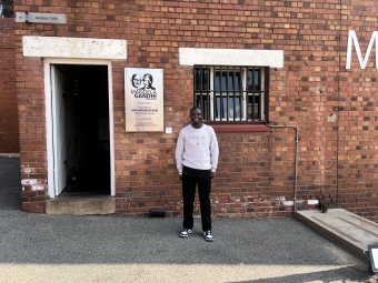 A man in a light grey sweatshirt and black pants stands in front of a red brick building at Constitution Hill in Johannesburg, next to an open doorway labeled "Isolation Cells" and a plaque commemorating the "Mandela Gandhi" exhibition.