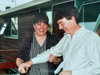 A laughing man and woman standing outdoors cut into a white frosted cake decorated with three strawberries.