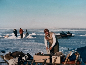 A man in a tan vest and striped hat smiles while organizing gear on a wooden sled on a vast, frozen expanse of sea ice.