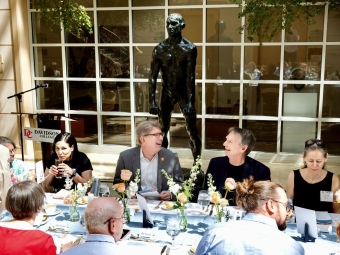 Two men sit at a decorated banquet table laughing together in front of a large bronze statue of a man and a windowed wall.
