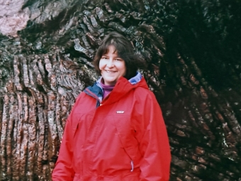 A smiling woman in a bright red hooded jacket stands in front of a textured, dark rock wall.