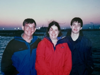 A man, woman, and teenage boy wearing outdoor jackets stand together smiling in front of a body of water at dusk.