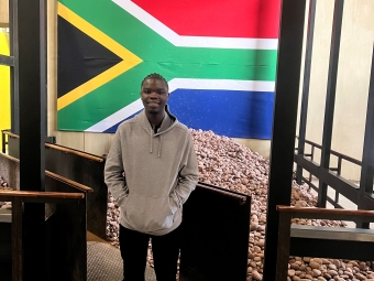 A man in a grey hoodie stands in an indoor exhibit featuring a large South African flag mounted above a pile of brown stones.