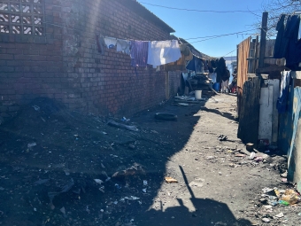 A narrow, sunlit dirt alleyway between a brick building with hanging laundry and a corrugated metal fence, showing signs of urban poverty.