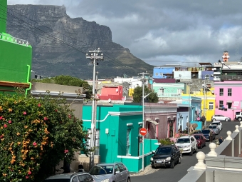A street view of the Bo-Kaap neighborhood in Cape Town, featuring brightly colored houses in green, pink, and teal with Table Mountain in the background under a cloudy sky.