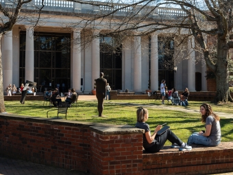 Students sit on a brick wall and at tables in a sunny campus courtyard featuring a large neoclassical building with white columns and an outdoor sculpture.