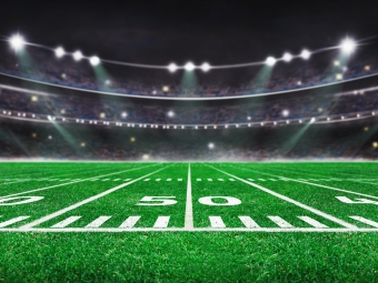 A wide-angle, low-altitude view of an American football field at night, focusing on the 50-yard line with a blurred stadium crowd and bright floodlights in the background.