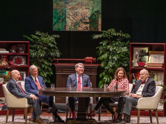 Five people sit around a large oval table on a stage set designed to look like a study, featuring bookshelves filled with memorabilia, potted plants, and an abstract painting hanging in the center.