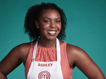 A smiling Black woman with curly hair wearing a white MasterChef apron with the name "ASHLEY" printed on it over a coral lace top, posed against a solid teal background.