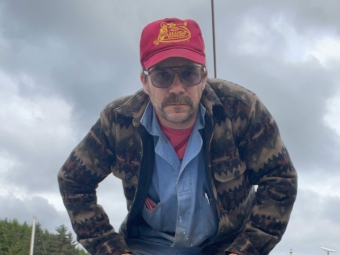 A man with a mustache, sunglasses, and a red Arkansas Razorbacks hat stands outdoors under a cloudy sky wearing a patterned wool jacket over a blue button-down shirt.