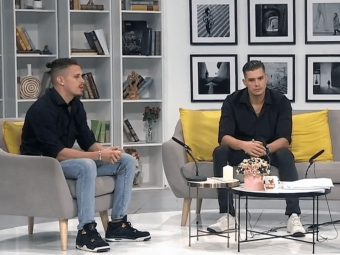 Two men in black shirts sit on a grey sofa with yellow pillows in a studio setting featuring bookshelves and framed wall art.