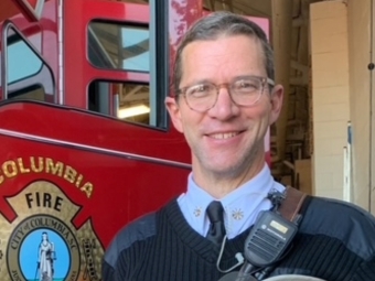 A smiling man in a black sweater and tie stands in front of a red fire truck featuring the "Columbia Fire" logo.