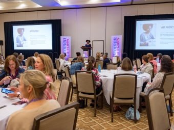 A presenter stands onstage, between two screens, in a hall filled with women