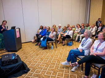A woman presents at a podium in a hall full of women