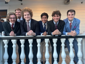 Six young men in formal suits and ties lean against a white balcony railing with classic balusters in front of a light-colored wooden house.