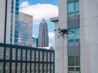 A professional-grade cleaning drone hovers beside a modern apartment building, spraying a stream of water onto the exterior windows against a city skyline featuring the Bank of America Corporate Center in Charlotte.