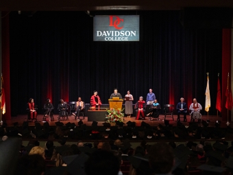 A wide view of a formal convocation ceremony on a stage with a "Davidson College" logo backdrop, featuring several faculty members in academic regalia seated around a student speaking at a podium