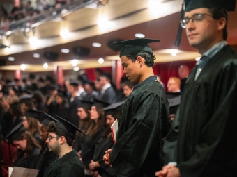 A side profile of a graduate in a black cap and gown standing with head bowed during a moment of reflection in a crowded auditorium.