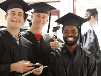 Three male students in black graduation caps and gowns smile for a close-up photo, with one student giving a thumbs-up.