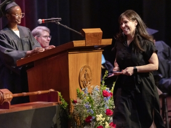 A woman in a black dress smiles as she walks across a stage with an award, while a student in academic regalia stands behind a wooden podium.