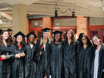 A group of eight smiling graduates in black academic gowns pose together indoors, with some holding red roses and diplomas.