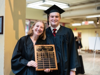 Two smiling graduates in black caps and gowns pose together while holding a wooden "Ben Callinder Award" plaque displaying several names.