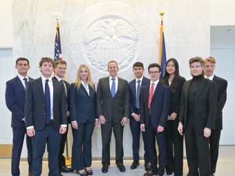 A group of ten professionally dressed students and a central official pose for a formal photo in front of a large, white architectural seal of the Federal Reserve Board of Governors.