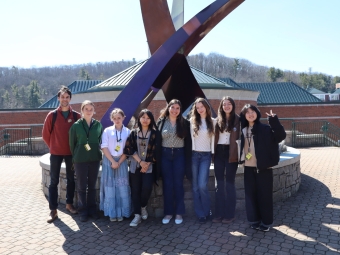 A group of eight students and a mentor pose outdoors in front of a large, dark abstract metal sculpture on a sunny day.