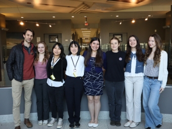 Seven young people stand side-by-side smiling in a brightly lit modern hallway with a decorative artistic ceiling.