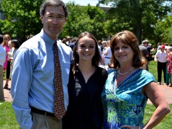 Three people at a graduation ceremony: a father in a tie, a graduate in a gown, and a mother in a blue dress