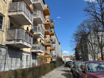 A sunlit city sidewalk lined with multi-story apartment buildings featuring rows of balconies on the left and parked cars along the street on the right under a blue sky with scattered clouds.