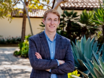 A young white man wearing a blue blazer in a tropical setting, arms crossed