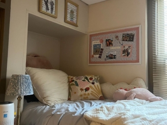 A bed with light grey bedding and a floral accent pillow is tucked into a wall nook, featuring a pink-bordered corkboard filled with photos and two gold-framed pictures above the headboard.