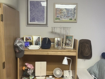 A wooden student desk with a hutch holds a collection of framed photos, jewelry, and vanity items, with two framed art pieces hanging on the wall above.