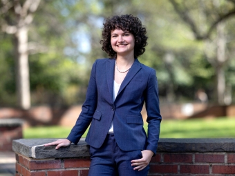 A woman with short curly hair smiles while wearing a navy blue suit and standing outdoors in front of a brick wall and blurred greenery.