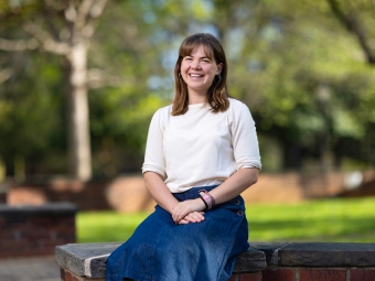 A smiling woman with shoulder-length brown hair and bangs sits on a stone-capped brick wall wearing a white top and blue denim skirt.