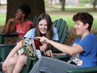 Two students sit in green Adirondack chairs outdoors, smiling as they look at a laptop screen covered in colorful stickers while a third student sits in the background.