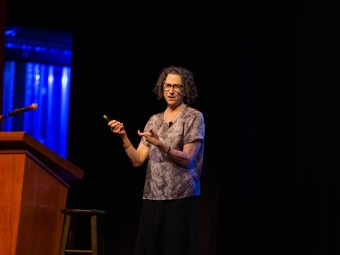 Dr. Cynthia Dwork gestures with a handheld clicker while delivering a lecture on a dimly lit stage next to a wooden podium.