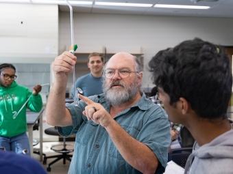Professor Durwin Striplin, a bearded man wearing glasses, points to a piece of glass laboratory equipment while instructing a student in a chemistry lab setting.