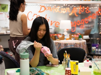 A student focused on her work uses a piping bag to apply white decorative frosting to a small heart-shaped pink jewelry box in a creative studio labeled "Davidson Arts & Creative Engagement."