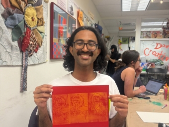 A smiling student with glasses and curly hair holds up a red piece of paper featuring three orange linocut prints of a face while sitting in a brightly decorated art studio.
