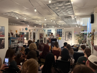An audience of students watches a live musical performance by a guitarist under warm string lights in a decorated community art space.