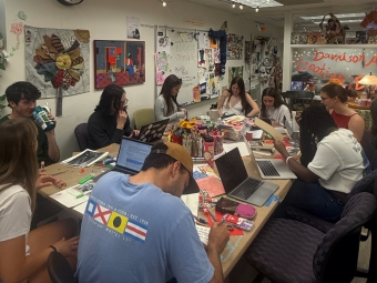 A group of students sits around a large, cluttered table in the Davidson Arts and Creative Engagement (DACE) Studio, working on various art projects and using laptops under warm string lights and a whiteboard filled with sketches.