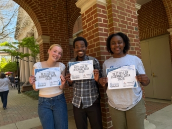 Three smiling students stand outside a brick building, each proudly holding a certificate that reads "Best Use of External Data 2026."