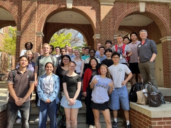 A large, diverse group of students and faculty pose together for a commemorative photo on the steps of a brick building with prominent arched entryways.