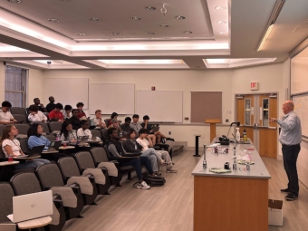 An instructor stands at the front of a tiered lecture hall, gesturing toward a whiteboard while a group of students sit at desks with their laptops.
