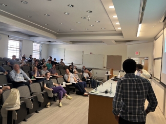 A student in a plaid shirt stands at the front of a classroom, presenting a data visualization slide featuring a pie chart to a seated audience of peers and judges.