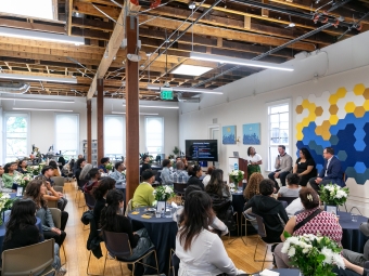 A seated audience watches a panel presentation in a modern, open-ceiling event space where four speakers are seated at the front next to a digital screen displaying a scholarship timeline.
